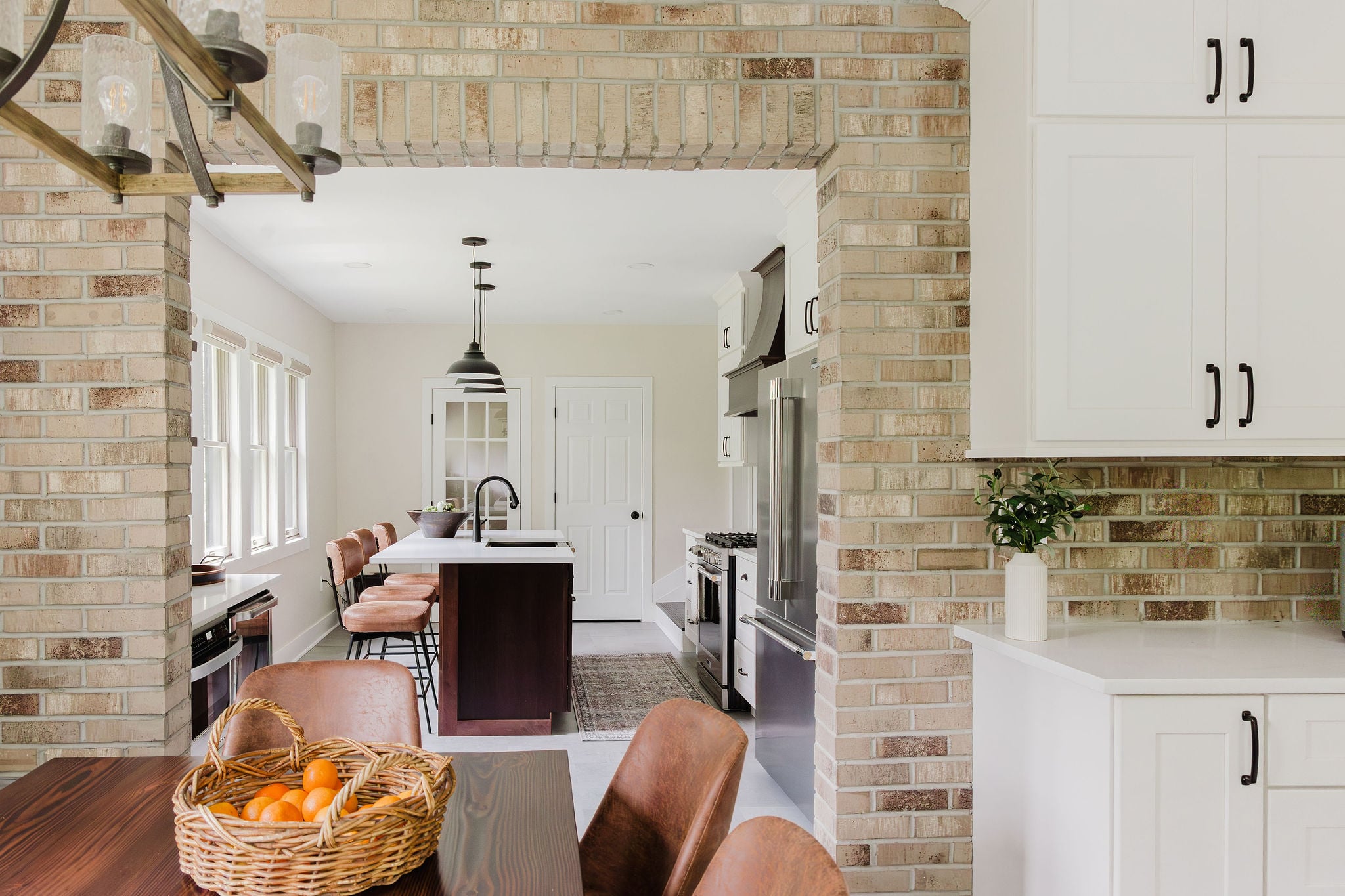 Brick archway looking into farmhouse kitchen with a dark hickory island, light grey concrete-look floor, black pendant lights, and cream white cabinets